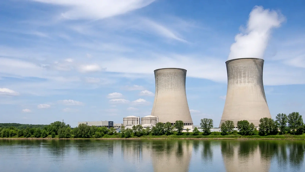 Two large cooling towers at a power plant along a calm river, with steam rising and a blue sky above.