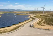 Repsol to Sell Renewables Portfolio Across Spain to Masdar Wide view of a solar farm with rows of solar panels and wind turbines on rolling hills under a blue sky.