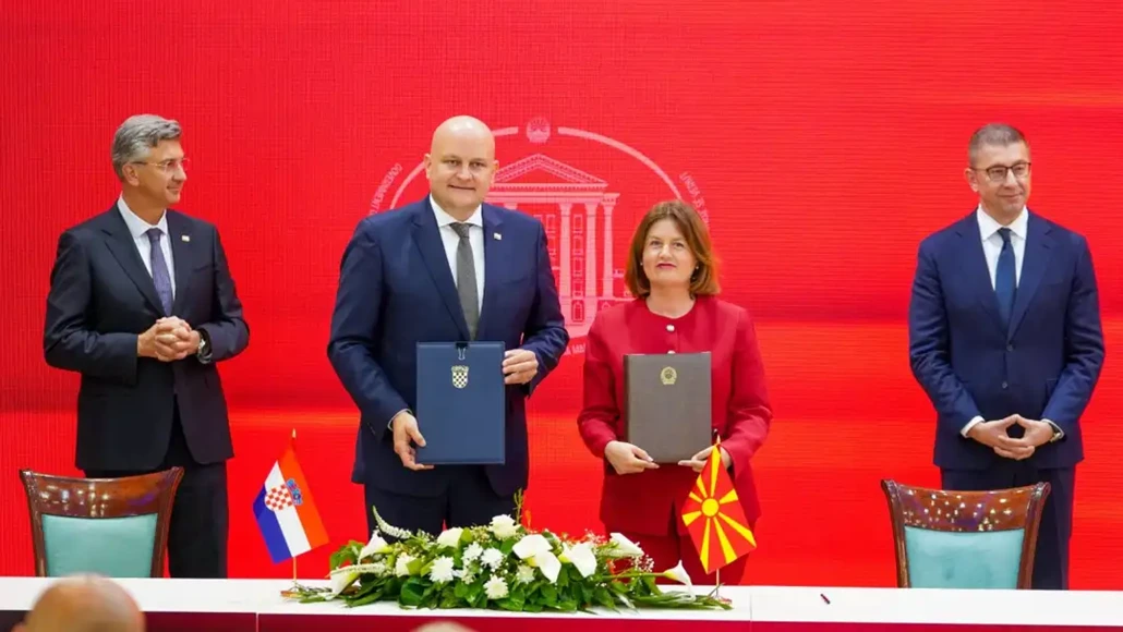 Officials at a signing ceremony holding blue and gray folders, with Croatian and North Macedonian flags on the table, against a red backdrop.