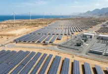 Somaliland Expands Renewables with Berbera Energy Project Coastal desert solar farm with rows of photovoltaic panels and wind turbines on the horizon near the sea and mountains.