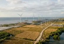 Meralco, Partners Sign 5GW Renewable Energy Project Deal Aerial view of a coastal wind farm with tall turbines, solar panels, and flat fields along the shoreline by the sea.
