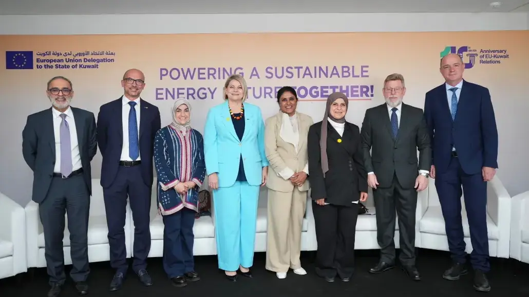 Group of eight professionals in formal attire posing in front of a banner reading “Powering a Sustainable Energy Future Together” at an EU–Kuwait relations event.