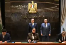 Egypt Signs Agreement to Build First Wind Turbine Factory Officials sign documents at a long desk as two men sit at the ends and a woman signs in the middle, with two men standing behind and a gold emblem on the wall behind them (official ceremony).