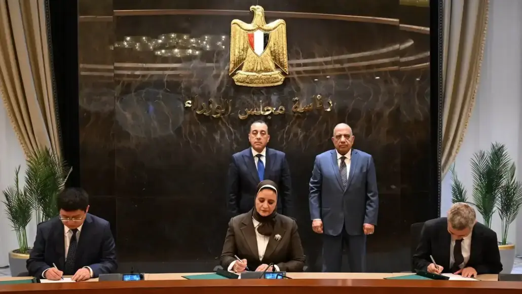 Officials sign documents at a long desk as two men sit at the ends and a woman signs in the middle, with two men standing behind and a gold emblem on the wall behind them (official ceremony).