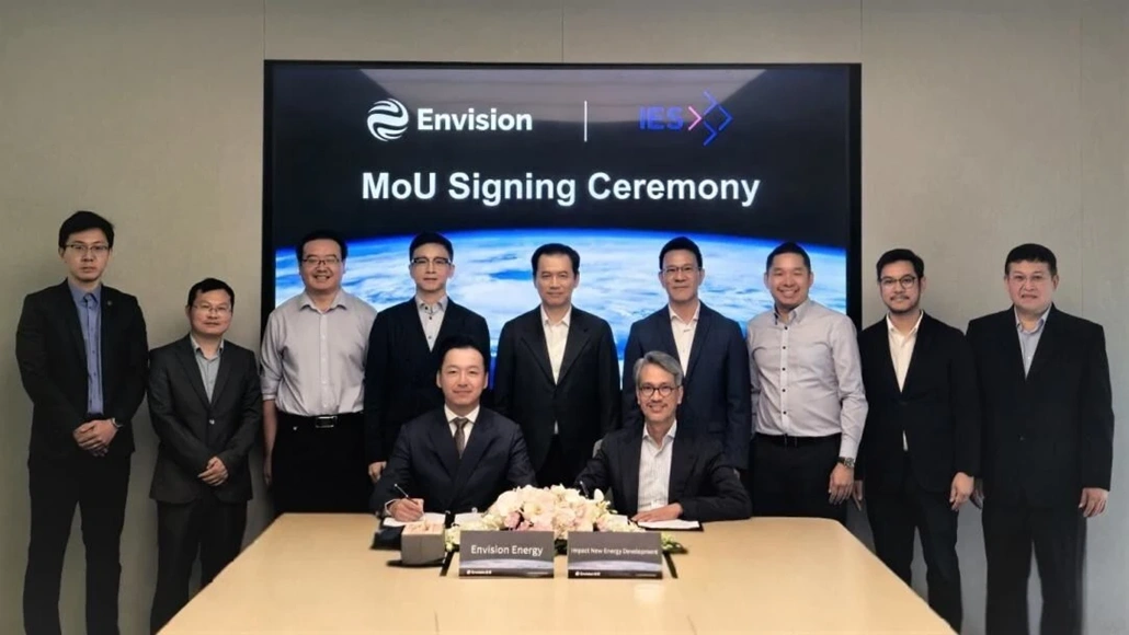 Two men sit at a conference table signing documents while nine colleagues stand behind them during a MoU signing ceremony, with a screen reading 'MoU Signing Ceremony' in the background.