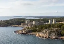 Rolls-Royce SMR Chosen for New Swedish Nuclear Power Project Industrial complex with cylindrical tanks and cooling towers on a rocky coastline, surrounded by trees and sea.