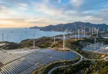 KOSPO Targets 11.2 GW Renewable Energy Capacity by 2040 Coastal renewable energy site with wind turbines on hills and solar panels in the foreground, the sea and mountains in the distance at sunset/ twilight.