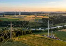 Germany Opens Electricity Grid Expansion Plan Consultation Aerial view of wind farm with tall turbines, power lines, and a river winding through green fields at sunset.