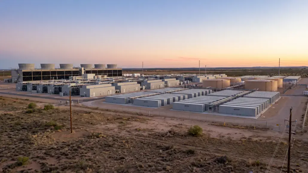Industrial complex in a desert landscape at sunset, featuring white rectangular buildings, row after row of modular units, and several large circular cooling towers in the background.