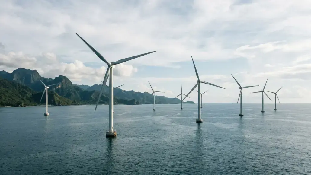 Offshore wind farm with multiple wind turbines standing in calm sea and a distant mountainous coastline behind them.