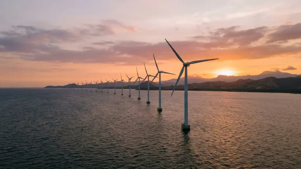 Offshore wind turbines lined up in the sea at sunset, with calm water and distant hills on the horizon.