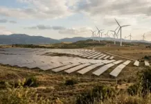 Turkey Launches New Renewable Energy Tender for Solar, Wind Rows of solar panels spread across a dry hillside with wind turbines on the distant horizon under a cloudy sky, showing renewable energy.