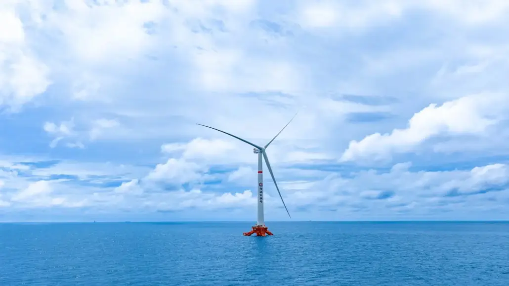 Offshore wind turbine standing in calm blue ocean under a partly cloudy sky, with three blades visible.