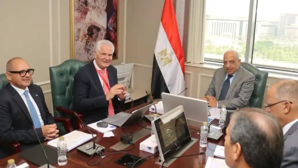 Group of five men in suits sit around a conference table with laptops, water bottles, and documents; a tall Egyptian flag stands by the window.