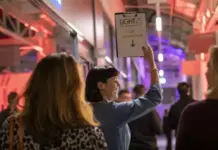 LiGHT 26 Registration Opens as Messe Frankfurt UK Launches the Next Chapter for the UK’s Lighting Specification Event Woman in a blue top raises a sign that reads 'LIGHT25 VIP DESIGN TOUR' in a busy, red-lit event hallway act of directing attendees.