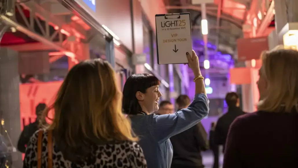 Woman in a blue top raises a sign that reads 'LIGHT25 VIP DESIGN TOUR' in a busy, red-lit event hallway act of directing attendees.