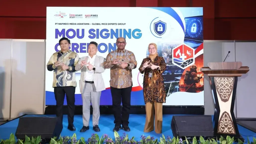 Four professionals on a blue-carpet stage posing with circular hand gestures in front of a banner that reads 'MOU SIGNING' ceremony behind them.