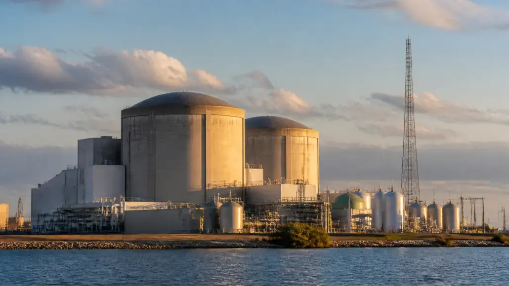 Nuclear power plant with two large cylindrical containment domes and cooling towers along a waterfront at sunset.