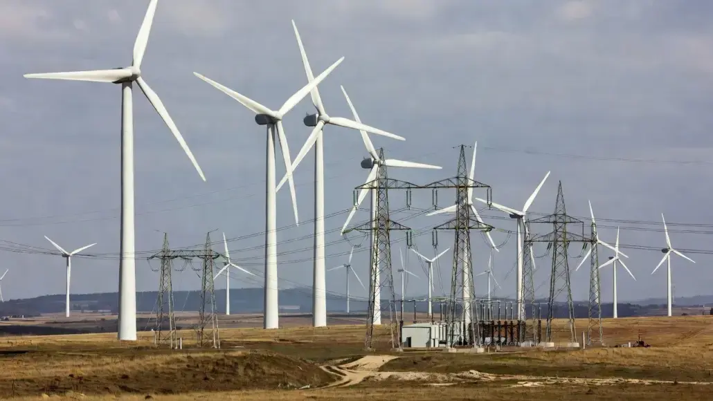 Wind farm with tall wind turbines and a nearby electrical substation in a flat, arid landscape.